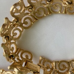 May include: A gold-toned bracelet with a detailed, intricate design. The bracelet is made of linked, open-work circles and has a clasp that reads "Monet".