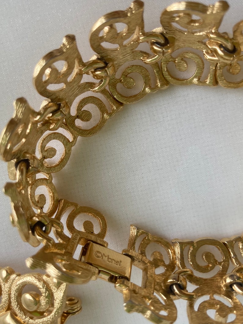 May include: A gold-toned bracelet with a detailed, intricate design. The bracelet is made of linked, open-work circles and has a clasp that reads "Monet".