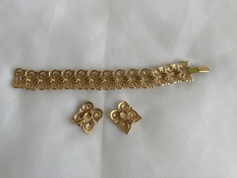 May include: A gold-tone bracelet and earrings set with a delicate, intricate design. The jewelry features a repeating pattern of swirls and loops.