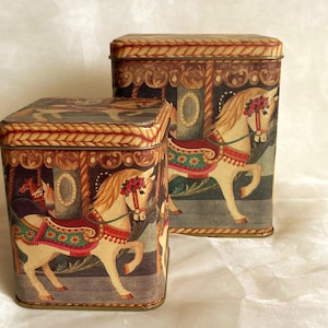 May include: Two rectangular metal storage tins with carousel horse designs. The tins feature a cream-coloured horse with a red and green saddle, a floral crown, and a carousel background. The tins have a decorative border and are set against a neutral background.
