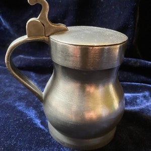 May include: A pewter tankard with a hinged lid and a handle. The tankard has a rounded body and a slightly flared base.