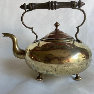 Op de afbeelding: Een vintage messing theepot met een houten handvat, een koperen deksel en drie poten. De theepot heeft een rond lichaam en een tuit.