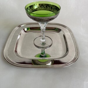 Peut inclure: Un verre à coupe teinté de vert avec un bord argenté et une tige torsadée repose sur un plateau carré argenté. Le verre présente un motif orné sur le bord. Le plateau a un bord arrondi.