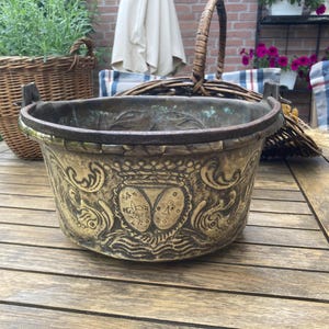 Può includere: Un'antica fioriera decorativa in metallo con disegni in rilievo ornati. La fioriera ha una forma ampia e rotonda con un bordo e manici scuri. Le sculture dettagliate includono motivi floreali ed araldici.