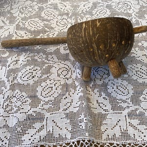Op de afbeelding: Een bruine kokosnootschelpzeef met een houten handvat en drie houten poten. De zeef staat op een wit gehaakt tafelkleed.