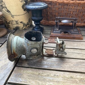 Op de afbeelding: Een antieke metalen lantaarn met een verweerde uitstraling. De lantaarn heeft een cilindrisch lichaam, een cirkelvormige lens en een handvat. Andere vintage items zijn een houten spoel, een decoratieve urn en een kleine metalen pers.