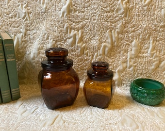 May include: Two amber glass jars with lids and a green glass bowl. The jars are square shaped and have a textured surface. The bowl is round and has a decorative design.
