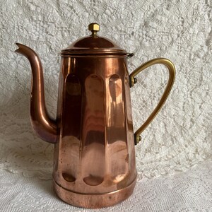 Peut inclure: Une cafetière en cuivre avec une poignée et un bouton de couvercle en laiton. La cafetière a un corps cylindrique et haut avec des indentations verticales et un bec courbé. La cafetière est présentée sur une nappe en dentelle blanche.