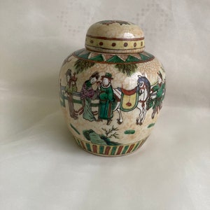Könnte beinhalten: Eine kleine, cremefarbene Keramikdose mit Deckel. Die Dose ist mit einer bunten Szene von Menschen und einem Pferd in einer Landschaft verziert. Die Dose hat einen grün-rot-braunen Streifenband um den Boden.