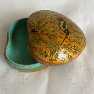 Op de afbeelding: Een hartvormige sieradendoos met een gouden buitenkant en een groen-gouden bloemenontwerp. Een kleine vogel is op de deksel geschilderd.