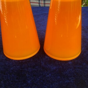 May include: Two orange glass cups with a smooth, rounded shape. The cups are sitting on a blue surface.