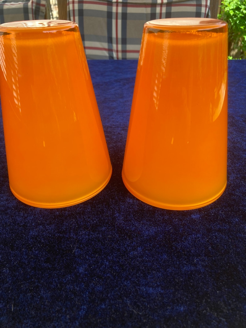 May include: Two orange glass cups with a smooth, rounded shape. The cups are sitting on a blue surface.