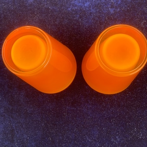 May include: Two orange glass candle holders with a frosted finish. The candle holders are empty and are sitting on a dark blue surface.