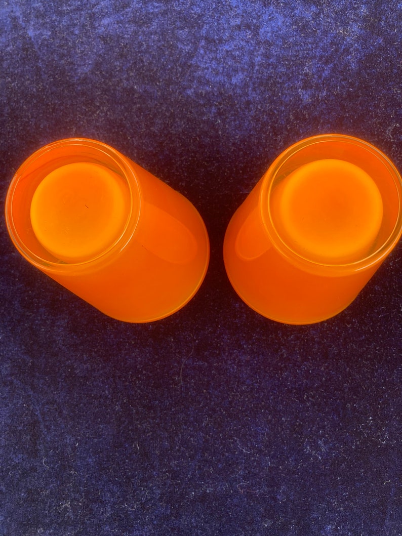 May include: Two orange glass candle holders with a frosted finish. The candle holders are empty and are sitting on a dark blue surface.