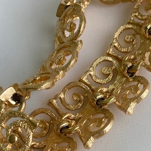 May include: Gold-tone bracelet with a delicate, intricate design featuring a repeating pattern of swirls and circles. The bracelet is made of metal and has a clasp closure.