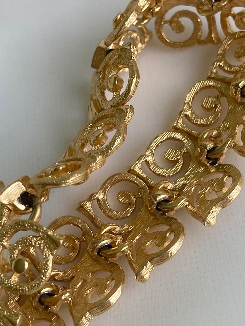 May include: Gold-tone bracelet with a delicate, intricate design featuring a repeating pattern of swirls and circles. The bracelet is made of metal and has a clasp closure.