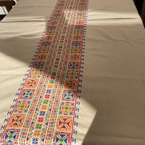 Könnte beinhalten: Weißer Tischläufer mit einem bunten bestickten Läufer mit geometrischen Mustern in Rot, Blau, Grün, Orange und Gelb.