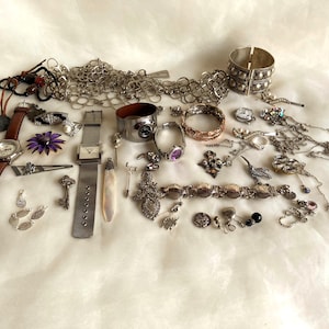 Op de afbeelding: Een verzameling vintage sieraden en accessoires, waaronder horloges met bruin lederen bandjes, zilveren kettingen, armbanden, ringen en oorbellen. Een sleutel, een paarse bloem en een manchetarmband zijn ook zichtbaar. De items zijn op een wit oppervlak geplaatst.