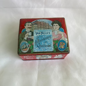 May include: A vintage tin box with a red, white and blue design. The box features an illustration of two people holding cups of tea. The text on the box reads "Van Nelle's Stoom Koffiebranderij en Theehandel".