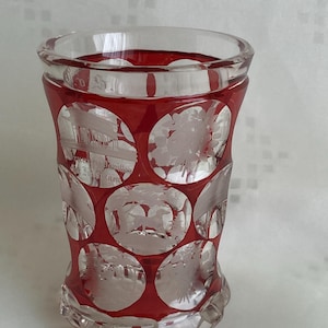 Può includere: Un bicchierino da shot in vetro rosso con un design inciso chiaro di cerchi con varie immagini. Il bicchiere ha un bordo smerlato e una base arrotondata.