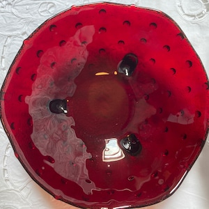 Può includere: Una ciotola di vetro rosso vibrante con una superficie strutturata e dettagli scuri. La ciotola ha una forma unica ed è adornata da piccoli abbellimenti scuri. Questo pezzo decorativo è ideale per servire o esporre.