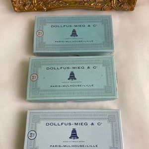Pode incluir: Três caixas vintage azuis com letras brancas e um logotipo de sino. As caixas são etiquetadas "Dollfus-Mieg & C" e "Paris-Mulhouse-Lille". As caixas são para fio de bordado DMC.