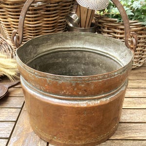 Op de afbeelding: Een cilindrische koperen emmer met een verweerde patina en een stevig, gebogen ijzeren handvat bevestigd aan twee zijlussen. Het metalen oppervlak vertoont tekenen van oxidatie en ouderdom, met een gladde, brede rand en een platte bodem.