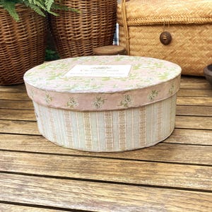 Pode incluir: Uma caixa de armazenamento redonda com tampa, apresentando uma tampa rosa claro com um padrão floral e uma etiqueta branca com o texto "Chapeau Fleur". As laterais da caixa são decoradas com listras verticais em azul claro, bege e branco.