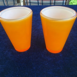 May include: Two orange glass cups with a white interior. The cups are sitting on a blue surface.