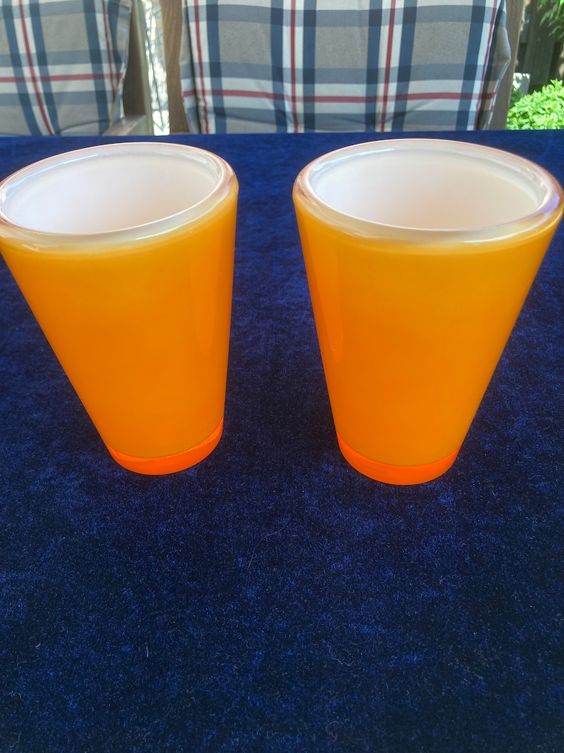 May include: Two orange glass cups with a white interior. The cups are sitting on a blue surface.