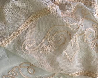 May include: A close-up of a white sheer fabric with delicate embroidered details in a cream colour. The fabric has a fine mesh texture and features intricate floral patterns.