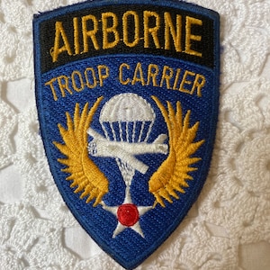 Op de afbeelding: Een blauw en gouden geborduurde patch met de woorden "Airborne Troop Carrier" en een witte parachute met vleugels.