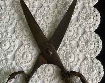 May include: A pair of antique scissors with a leather handle on one side and a metal ring on the other. The scissors are lying on a white crocheted doily.