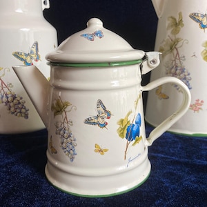 Puede incluir: Tetera de esmalte blanco con borde verde, adornada con ilustraciones de mariposas y uvas. La tetera tiene tapa y asa. Otros utensilios de cocina a juego son visibles en el fondo.