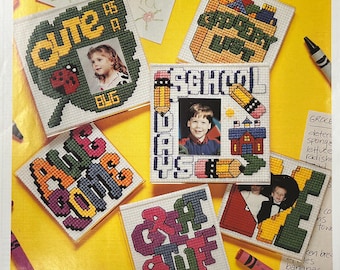May include: Six colorful cross-stitch designs on a white background with a clear acrylic frame. The designs include "Cute as a Bug", "School Days", "Grocery List", "All Aboard", "Craft Stuff", and "Home Sweet Home". The designs are framed with a clear acrylic magnet.