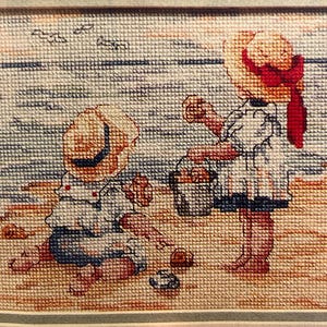May include: Cross-stitch embroidery of two children playing on a beach. One child is wearing a blue and white outfit and a straw hat, while the other child is wearing a white dress and a red hat. The beach scene is depicted with blue waves and sand.