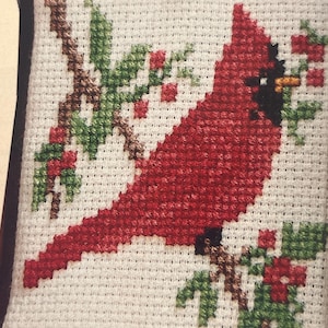 May include: Cross-stitch design of a red cardinal bird perched on a brown branch with green leaves and red berries. The bird has a black beak and eye. The background is white with red stitching along the top and bottom edges.