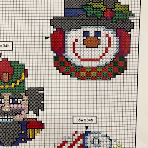 May include: Cross-stitch pattern featuring a snowman wearing a hat and earmuffs, with a wreath of green leaves and red berries. The pattern is 30 squares wide by 31 squares high. The design is on a grid, ready for stitching.