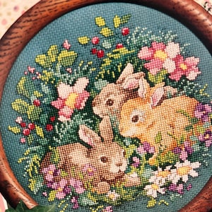 Może przedstawiać: Haft krzyżykowy przedstawiający trzy króliki pośród kwiatów i liści. Projekt zawiera różowe, białe i fioletowe kwiaty, z czerwonymi jagodami i zielonymi liśćmi. Dzieło sztuki jest osadzone w okrągłej drewnianej ramie.