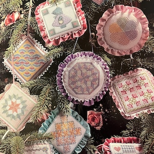 Könnte beinhalten: Neun handgefertigte Weihnachtsornamente mit verschiedenen Designs, darunter ein Stern, ein Baum, ein Herz und ein geometrisches Muster. Die Ornamente sind alle aus weißem Stoff gefertigt und haben rosa, lila, blaue und grüne Akzente. Sie hängen an einem grünen Weihnachtsbaum.