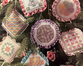 May include: Nine handmade Christmas ornaments with various designs, including a star, a tree, a heart, and a geometric pattern. The ornaments are all made with white fabric and have pink, purple, blue, and green accents. They are hanging on a green Christmas tree.