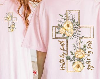 May include: Light pink t-shirts with a gold cross design. The front of the shirt has a small cross with yellow flowers. The back of the shirt has a larger cross with yellow flowers and the text "Walk by faith, not by sight."