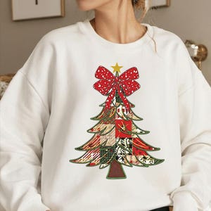 May include: White sweatshirt featuring a Christmas tree design. The tree is composed of various patterned patches in red, green, and gold, topped with a red bow and a yellow star. The design is centered on the front of the sweatshirt.