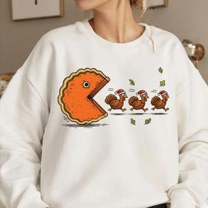 May include: White sweatshirt featuring a cartoon pie character chasing three turkeys wearing Santa hats. The pie has a large bite taken out of it, resembling a Pac-Man design. Green leaves are scattered around the turkeys.