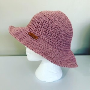 May include: A pink crocheted sun hat with a brown label. The hat is on a white mannequin head.