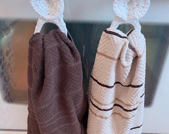 May include: Two kitchen towels hanging from a white crocheted loop. One towel is a solid dark brown with a textured pattern. The other is off-white with brown stripes and a chevron pattern. The crocheted loops are attached to a white surface.