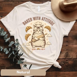 May include: A natural-colored t-shirt featuring a cartoon cat wearing a chef's hat and holding a rolling pin. The text "BAKED WITH ATTITUDE" is arched above the cat, with croissants and clouds as design elements.