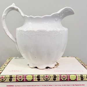 May include: A white ceramic pitcher with a scalloped rim, a curved handle, and a textured lace-like pattern around the upper body. The pitcher features a rounded, fluted base and a pouring spout, displaying a glossy finish throughout the surface.