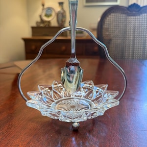 Op de afbeelding: Een vintage suikerpot met zilveren coating en een heldere glazen schaal. De schaal heeft een sterrenpatroon en een zilveren lepel. De suikerpot staat op een houten tafel.