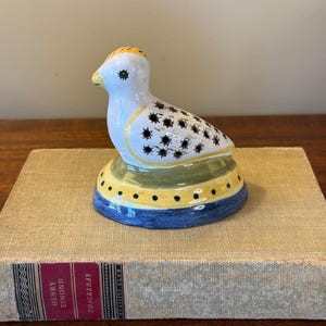 May include: A small ceramic bird figurine with black and white polka dots, yellow accents, and green and blue details. The bird is sitting on a base with a yellow and blue design.